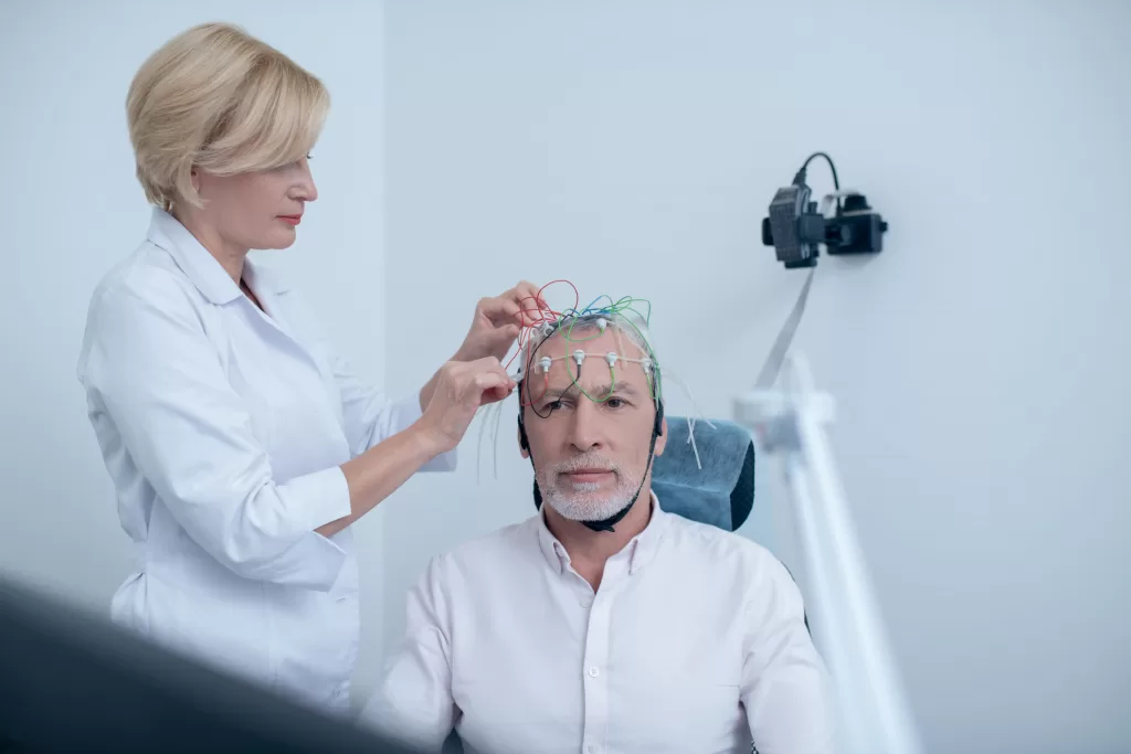 Female neurologist adjusting electrodes on gray-haired male patient head undergoing electroencephalogram