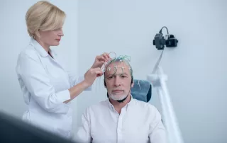 Female neurologist adjusting electrodes on gray-haired male patient head undergoing electroencephalogram