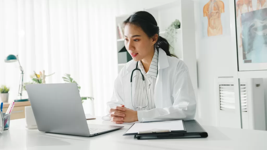 Young female doctor in white medical uniform with stethoscope using laptop for online consultation with a patient